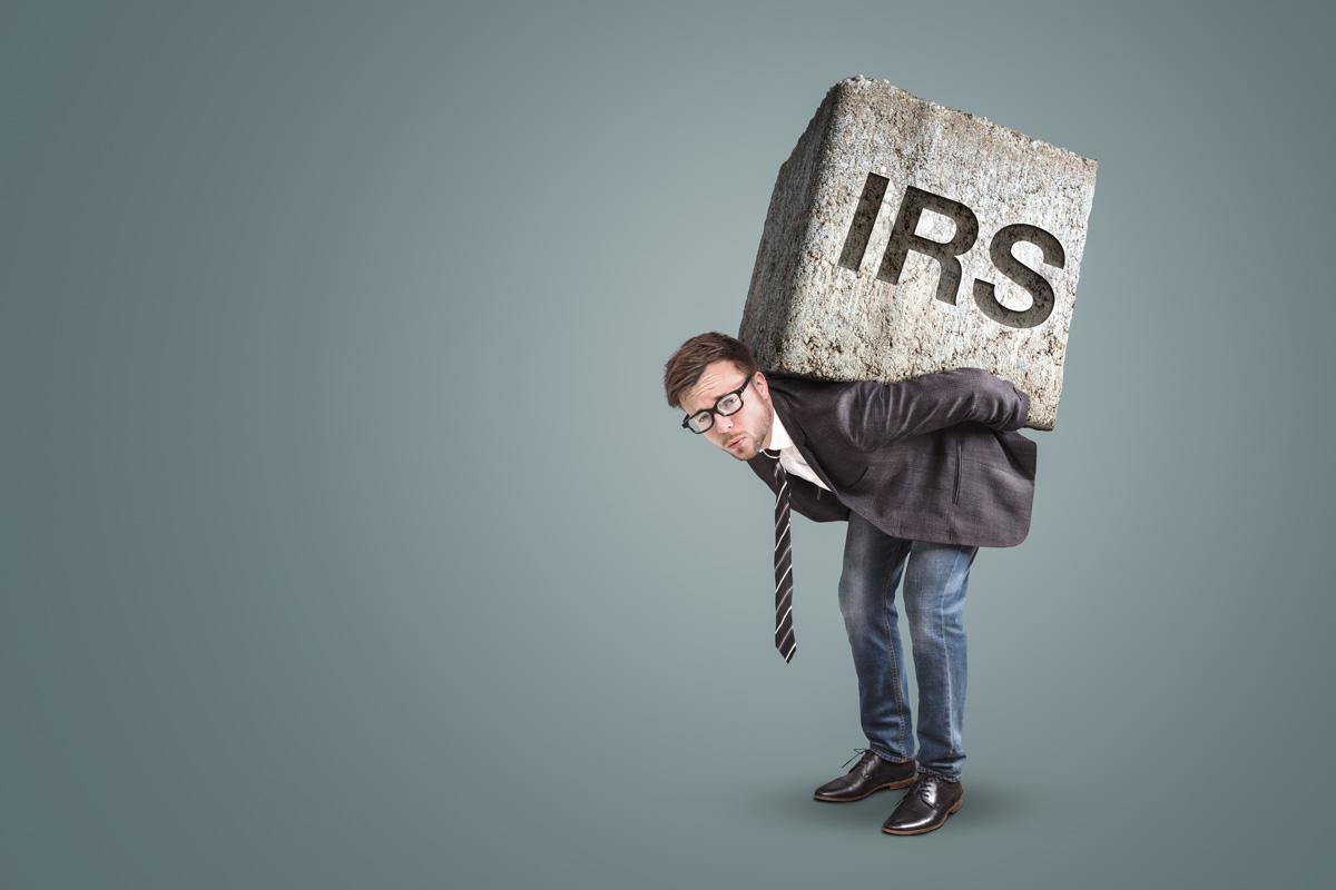 A person in a suit carrying a cement block on their back that reads “IRS” in El Paso.